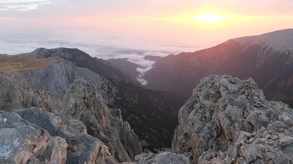 Θέα από την κορυφή Μύτικας στον Όλυμπο, με βράχια και τον ήλιο κοντά στον ορίζοντα.