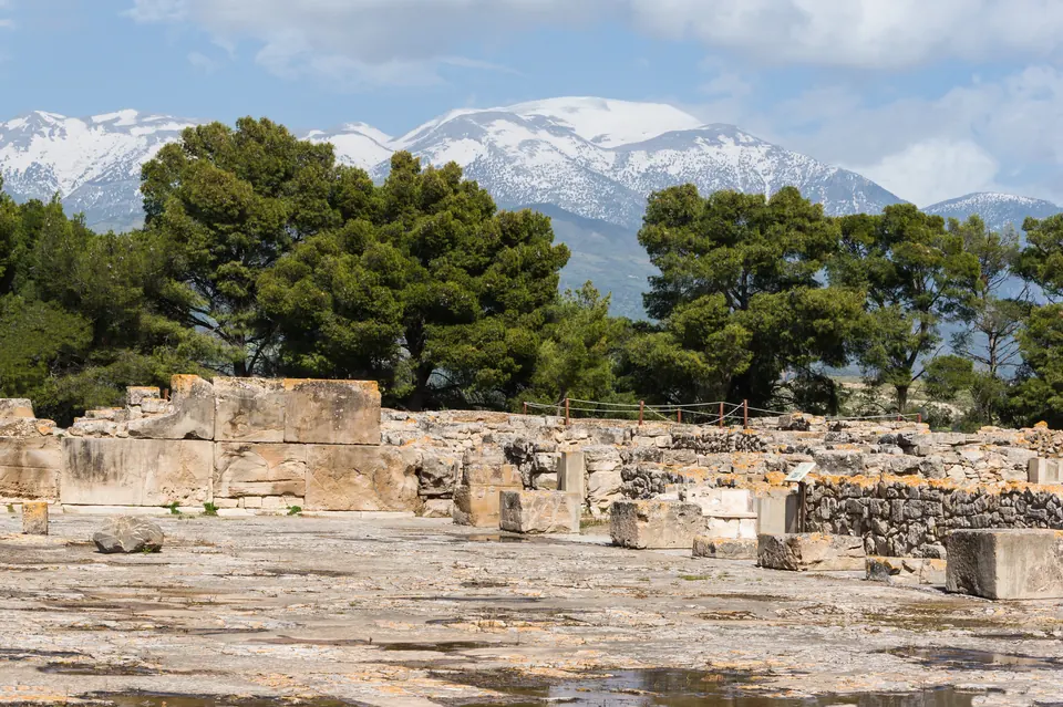 Η κεντρική αυλή του ανακτόρου της Φαιστού στην Κρήτη, με αρχαία θεμέλια και χιονισμένες κορυφές πίσω της.