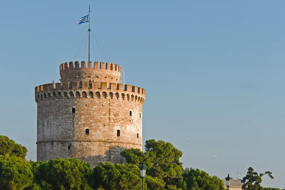 Ο Λευκός Πύργος της Θεσσαλονίκης με ελληνική σημαία, πίσω από πεύκα, κάτω από καθαρό γαλάζιο ουρανό.