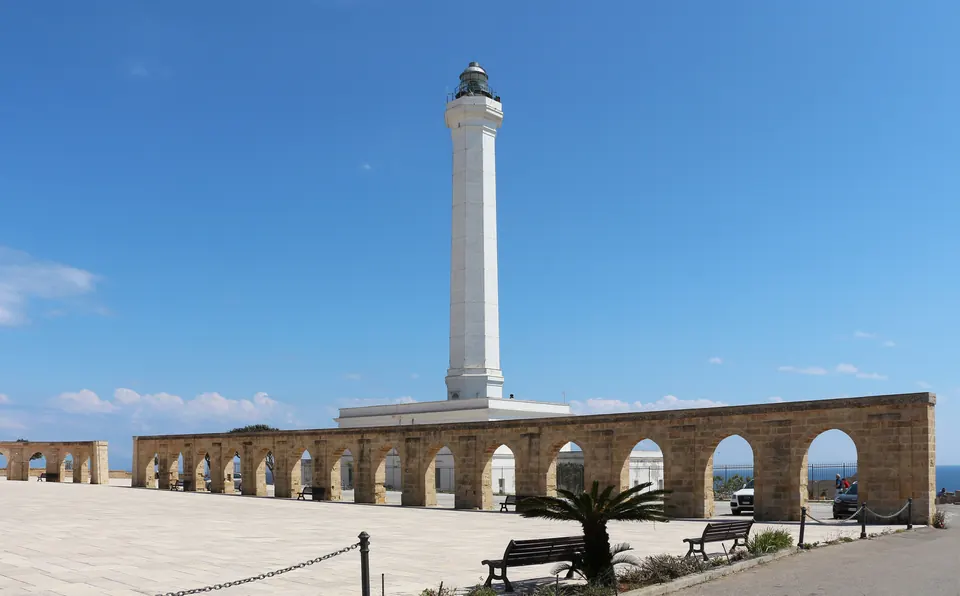 Ο φάρος του Capo Santa Maria di Leuca και μια πέτρινη στοά με αψίδες, κάτω από γαλάζιο ουρανό.