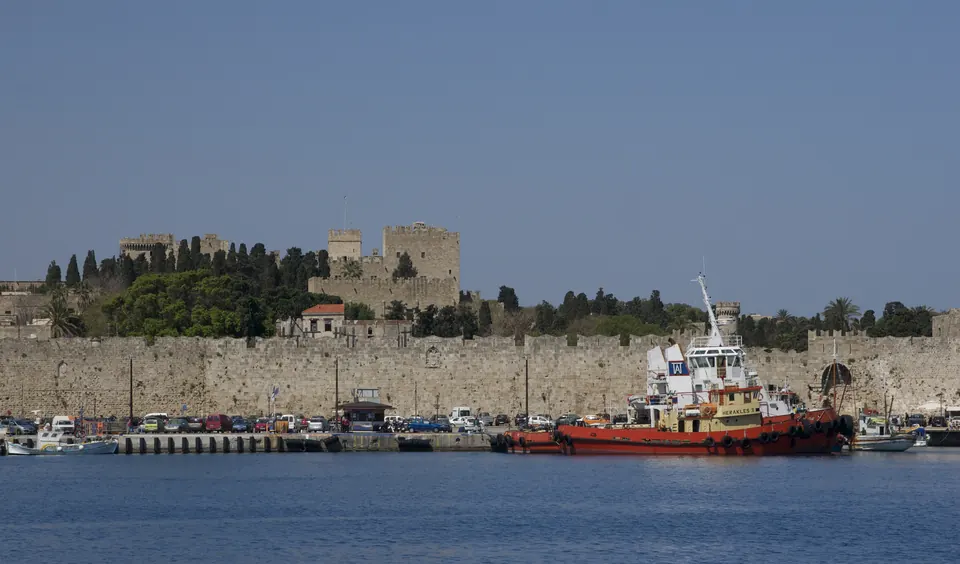 Τα τείχη της παλιάς πόλης της Ρόδου με το Παλάτι του Μεγάλου Μαγίστρου πίσω τους και ένα ρυμουλκό στο λιμάνι.